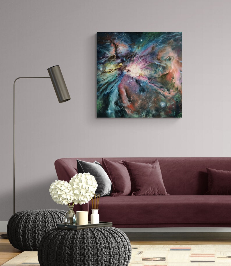 May include: A painting of a nebula in shades of blue, purple, pink, and green. The nebula is surrounded by a dark blue background with white stars. The painting is framed and hanging on a wall above a burgundy sofa.