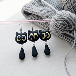 May include: Three pairs of black cat-shaped earrings with large, round, cream-colored eyes and teardrop-shaped dangles. The earrings are hanging on a pink rod, with gray yarn and knitting needles in the background.