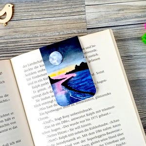 Peut inclure: Marque-page aquarelle représentant un paysage côtier au clair de lune avec pleine lune, eau et rivage. Le marque-page est dans un livre ouvert, avec des découpes d'oiseaux en bois et des stylos.