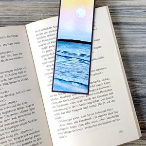 Peut inclure: Marque-page aquarelle représentant un paysage avec une pleine lune dans un ciel jaune et rose, au-dessus d'un océan bleu et blanc. Le marque-page est rectangulaire avec une bordure violet foncé et est placé dans un livre ouvert.