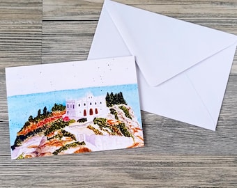 May include: A watercolor greeting card featuring a coastal scene with a white building on a rocky cliff, blue water, and green trees. A white envelope is next to the card. The card has a light blue sky.