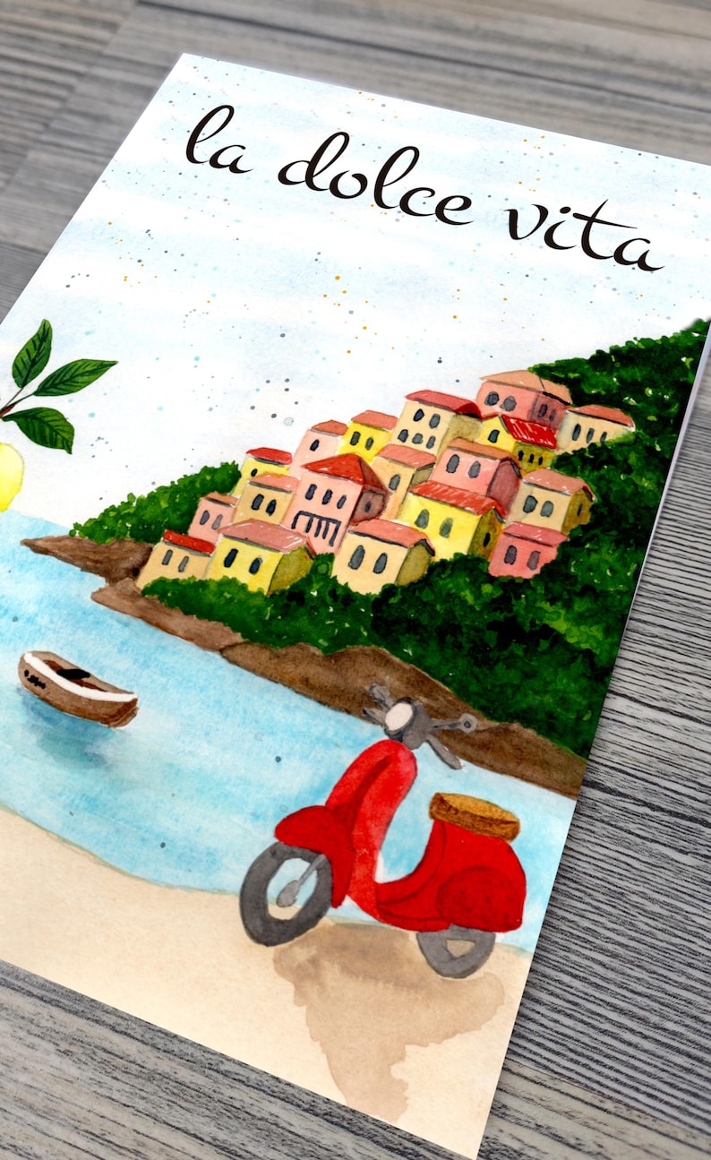 May include: Watercolor painting of a coastal Italian village with colorful buildings and a red scooter. The phrase "la dolce vita" is written in cursive at the top. A boat floats in the blue water.