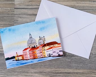 May include: Watercolor painting of a cityscape with domes and buildings in shades of red, orange, and yellow. The painting is on a card with a white envelope. The card depicts a waterfront scene with a blue sky and water.