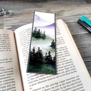 Peut inclure: Marque-page aquarelle représentant une forêt de conifères et une chaîne de montagnes dans des tons de vert, de violet et de blanc. Le marque-page est rectangulaire avec un bord gris foncé et est posé sur un livre ouvert.