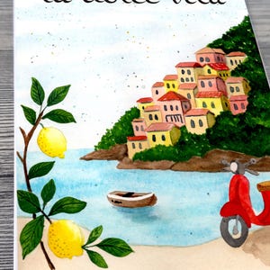May include: Watercolor illustration featuring the phrase "la dolce vita" above a coastal scene. The image depicts colorful buildings on a hillside, a red scooter, a small boat, and lemon branches.