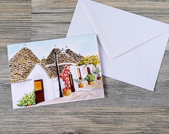 May include: A watercolor greeting card featuring a quaint village scene with white buildings, cone-shaped roofs, and a street lamp. The card is accompanied by a white envelope, perfect for sending a personalized message. The artwork showcases a charming European town.