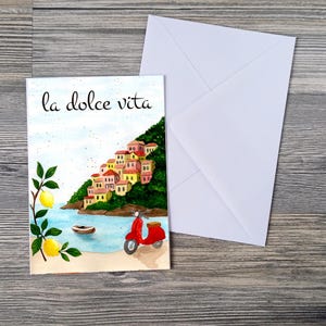 May include: Watercolor greeting card with the phrase "la dolce vita" and a coastal Italian scene. The artwork features a red scooter, a small boat, and a hillside village with colorful buildings. A white envelope is next to the card.