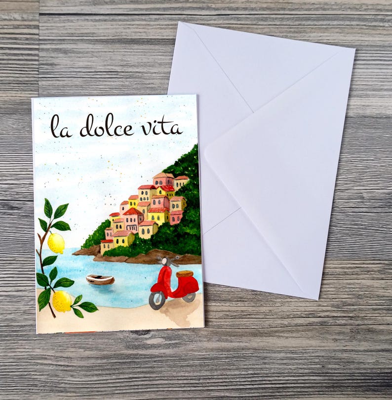 May include: Watercolor greeting card with the phrase "la dolce vita" and a coastal Italian scene. The artwork features a red scooter, a small boat, and a hillside village with colorful buildings. A white envelope is next to the card.