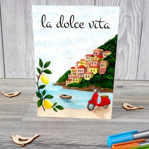 May include: Watercolour art print with the words "la dolce vita." The image shows a coastal Italian scene with colourful buildings on a hillside, a red scooter, lemons, and a small boat on the water.