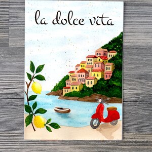 May include: Watercolor illustration of a coastal Italian scene with the text "la dolce vita." The image features a red scooter, buildings on a hillside, a boat, lemons, and the sea.
