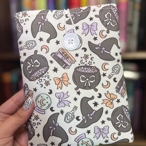 May include: A white book sleeve with a repeating pattern of witch hats, cauldrons, bows, moons, stars, and potion bottles in gray, orange, and purple. The sleeve is secured with a white button and elastic loop.