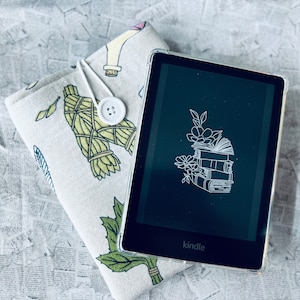 May include: A black Kindle e-reader with a white outline of a stack of books and flowers on the screen. The e-reader is on top of a white and green fabric with a floral pattern.