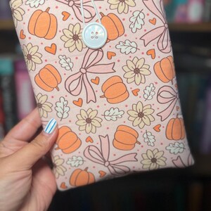 May include: A fabric book sleeve featuring a repeating pattern of orange pumpkins, pink bows, and floral elements on a light pink background. The sleeve is secured with a white elastic loop and a light blue button. The design evokes a fall or autumn theme.