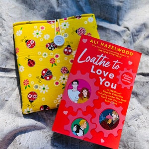 May include: A red and pink paperback book titled "Loathe to Love You" by Ali Hazelwood. The book cover features a cartoon illustration of a couple and a gear design. The book is sitting on a yellow fabric book cover with a ladybug pattern.