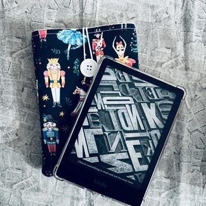 May include: A black and white Kindle e-reader with a black and white cover featuring a Nutcracker theme. The cover has a white button and the text "Choose something to read and see its cover here" and "Kindle" are visible on the screen.