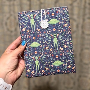 May include: A navy blue book cover with a repeating pattern of green aliens, flying saucers, moons, stars, and paisley shapes. The cover is secured with a white button and elastic loop. The overall theme is space and extraterrestrial.