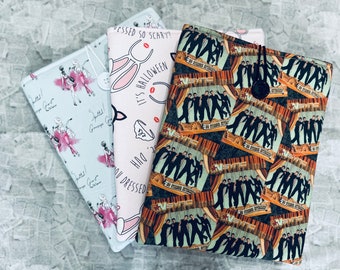 May include: Three different fabric covers for a tablet or e-reader. The first cover is light blue with a white and pink pattern of women in dresses. The second cover is pink with a white bunny rabbit and the text "It's Halloween!  You dressed so scary?  Duh!" The third cover is black with a white and brown pattern of a band with the text "NSYNC" repeated.