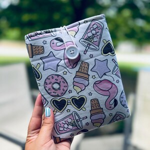 May include: A light gray fabric book cover with a repeating pattern of pink flamingos, ice cream cones, sunglasses, donuts, and stars. The cover is secured with a white string and a large, round button.