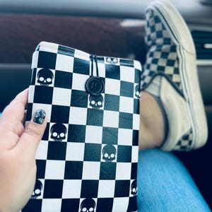 May include: Black and white checkered book cover with skull graphics. The cover is secured with a black button and string closure. The design features a repeating pattern of skulls within the checkered squares. The cover is held in a hand.