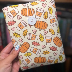 May include: Cream-colored book sleeve featuring a repeating pattern of orange pumpkins, coffee cups, autumn leaves, and pink hearts. The sleeve has a white button closure and a white elastic loop. The background is a blurred bookshelf.
