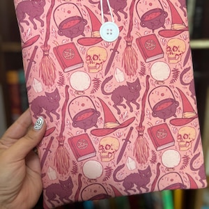 May include: A pink book cover with a repeating pattern of witch-themed illustrations. The design includes black cats, cauldrons, witch hats, brooms, skulls, and spell books. A light blue button and string closure is visible.