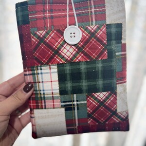May include: A patchwork book cover featuring a variety of plaid patterns in red, green, and cream. The cover is secured with a white button and elastic loop closure. The design is a mix of different plaid fabrics.