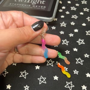 May include: A collection of colorful gummy worm earrings. One earring is held up by a hand, showing a pink and blue worm. Two other earrings are visible, one green and orange, and the other orange and yellow. The earrings are on a black surface with white stars.