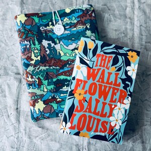 May include: A blue and green book cover with a floral design and the title "The Wall Flower Sally Louise" in red and white letters. The book is sitting on top of a blue and green patterned fabric book cover.