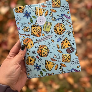 May include: A light blue book cover with a repeating pattern of yellow dice, stars, flowers, and other game-related graphics. The cover is secured with a white button and string.