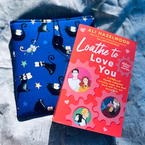 May include: A red paperback book titled "Loathe to Love You" by Ali Hazelwood. The book cover features a gear design with a couple in the center. The book is a STEM novella collection including "Under One Roof", "Stuck with You", and "Below Zero".