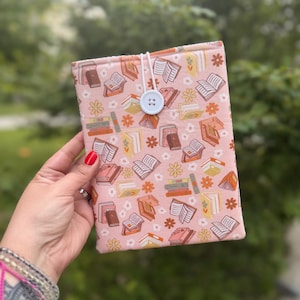 May include: A pink fabric book cover featuring a repeating pattern of open and closed books, and flowers in shades of orange, brown, and white. The cover has a light blue button closure and a white string. The cover is designed to protect a book.