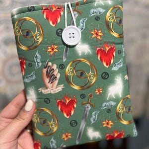 May include: Green fabric book sleeve with a white button closure. The sleeve features a repeating pattern of gold-colored ornate circles, swords, flaming hearts, and a hand with long black nails. The text "Love Within" is also visible.