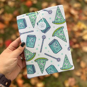 May include: White book cover with a repeating pattern of green witch hats, spell books, and wands. The book cover is secured with a white button and string closure. The spell books have the word "Spells" printed on them.