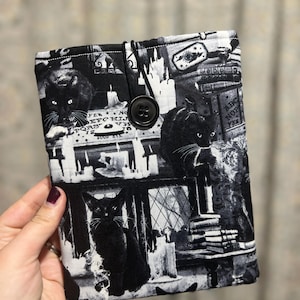 May include: A black and white fabric book sleeve featuring a repeating pattern of black cats, candles, and gothic architectural elements. The sleeve has a button closure and a black elastic loop. The overall theme is spooky and gothic.