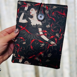 May include: A black book sleeve featuring a gothic illustration of a woman in a black dress with red ribbons. The sleeve has a black button closure and a black elastic loop. The background is a dark floral pattern.