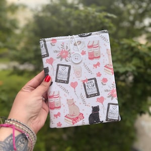 May include: White fabric book cover with a pattern of pink hearts, cats, coffee cups, books, and e-readers. The cover is secured with a white button and a loop closure. The background is a blurred green.