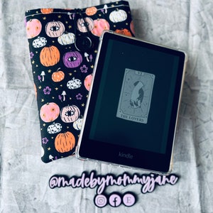 May include: A black and white patterned fabric pouch with a drawstring closure. The pouch is decorated with pumpkins, stars, and eyes. A black Kindle e-reader is inside the pouch. The screen of the Kindle displays the Lovers card from a tarot deck.