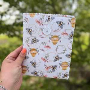 May include: A white book cover with a bee and honeycomb pattern. The design includes black bees, yellow honeycombs, and pink floral accents. A white button and elastic closure are visible.