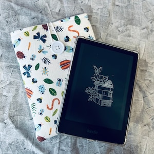 May include: A white and black Kindle e-reader with a black and white illustration of a stack of books and flowers on the screen. The Kindle is sitting on top of a white fabric case with a colorful insect and leaf pattern. The case has a button on the front.