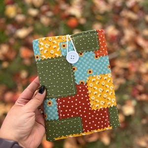 May include: A patchwork fabric book cover with a button closure. The cover features a mix of green, yellow, blue, and red fabric squares with floral patterns. A white button and string secure the cover.