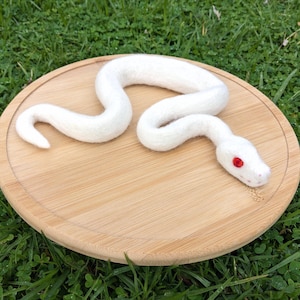 May include: A white felt snake with red eyes, curled up on a wooden surface. The snake is made of wool and has a realistic appearance.