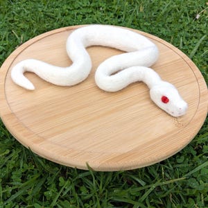 May include: A white felt snake with red eyes, curled up on a round wooden surface.