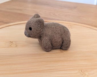 May include: A handmade felt wombat figure in shades of brown. The small, rounded animal has a dark grey nose and small, black eyes. It is posed on a light brown wooden surface, showcasing the handmade details of the wool sculpture.