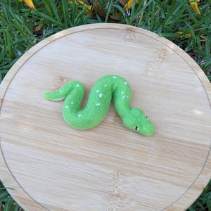 Puede incluir: Una serpiente de fieltro verde con manchas blancas, enroscada sobre una superficie de madera.