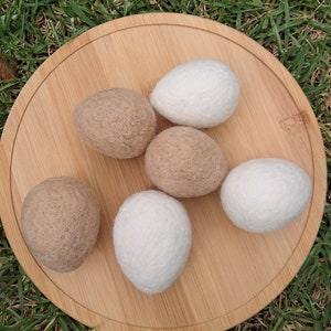 May include: Six felted wool balls, three white and three brown, arranged on a wooden surface. The balls are round and have a soft, fuzzy texture.