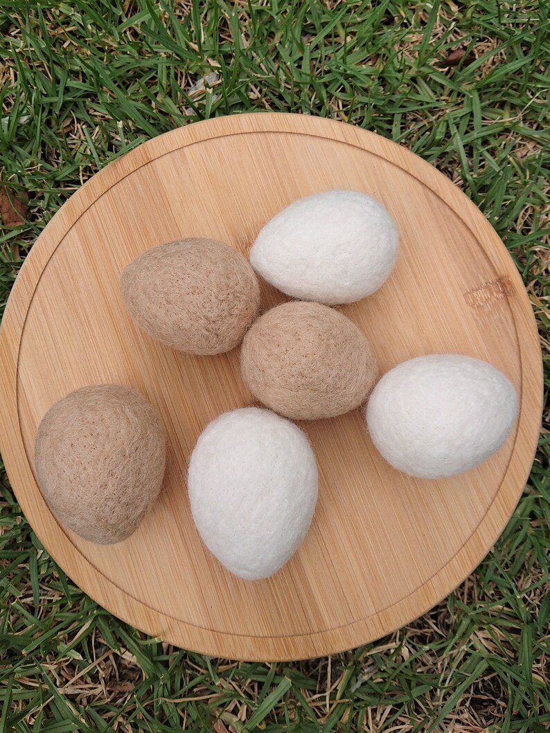 May include: Six felted wool balls, three white and three brown, arranged on a wooden surface. The balls are round and have a soft, fuzzy texture.
