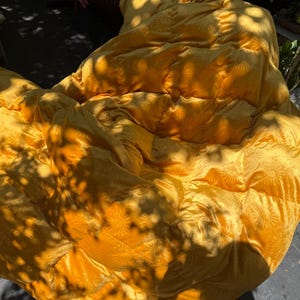 May include: A large, golden-yellow quilted blanket with a sunburst pattern. The blanket is folded and appears soft and plush. The sunlight casts shadows across the surface, creating a textured effect.
