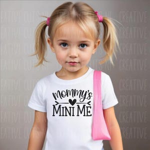 May include: A young girl with blonde pigtails wearing a white t-shirt printed with "Mommy's Mini Me" in black. She has a pink strap over her shoulder. The background is a light grey.