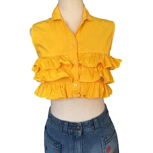May include: A vibrant yellow sleeveless top with a collared neckline and button closure. The top has multiple layers of ruffled fabric across the chest. It is worn with blue denim jeans.