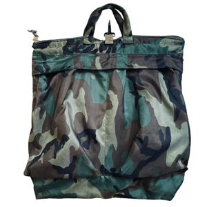 Puede incluir: Un bolso tote de camuflaje con asa superior y bolsillo delantero. El bolso presenta un cierre de cremallera y un patrón de camuflaje verde, marrón y negro. El bolso tiene forma rectangular y una construcción robusta.
